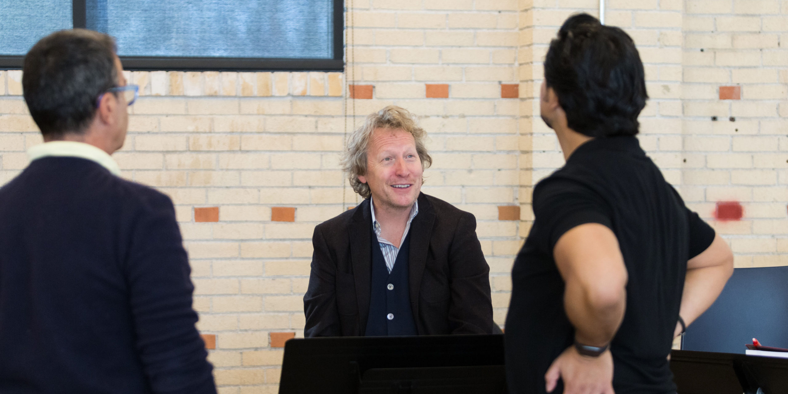 Ari Pelto smiles, facing the camera, while conversing with two cast members who have their backs to the camera.