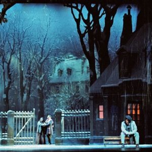 Snowy trees surround the set. A home with warm glowing windows sits to the right of the stage. Two lovers embrace center stage next to a gate.
