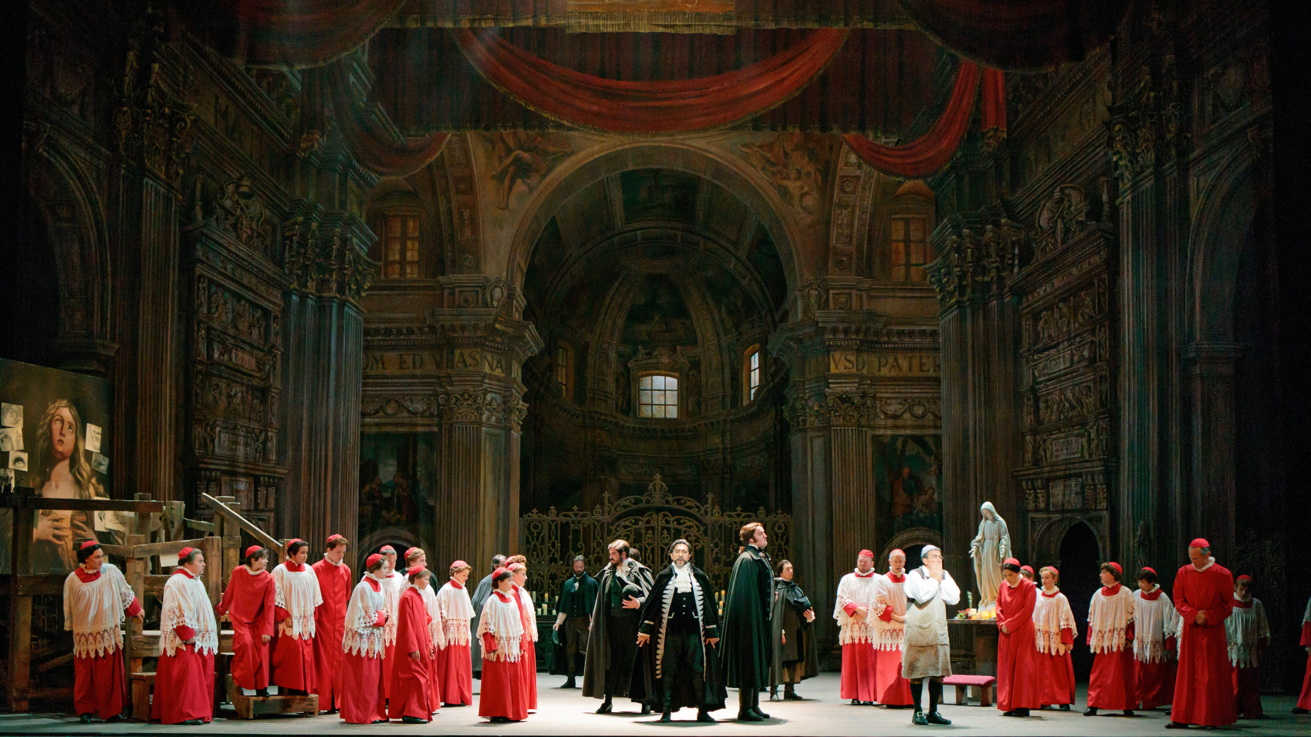 Baritone Luis Ledesma stand powerfully at center stage, flanks on either side by choir boys in red church robes. The set depicts the grand Sant’Andrea della Valle cathedral in Rome.