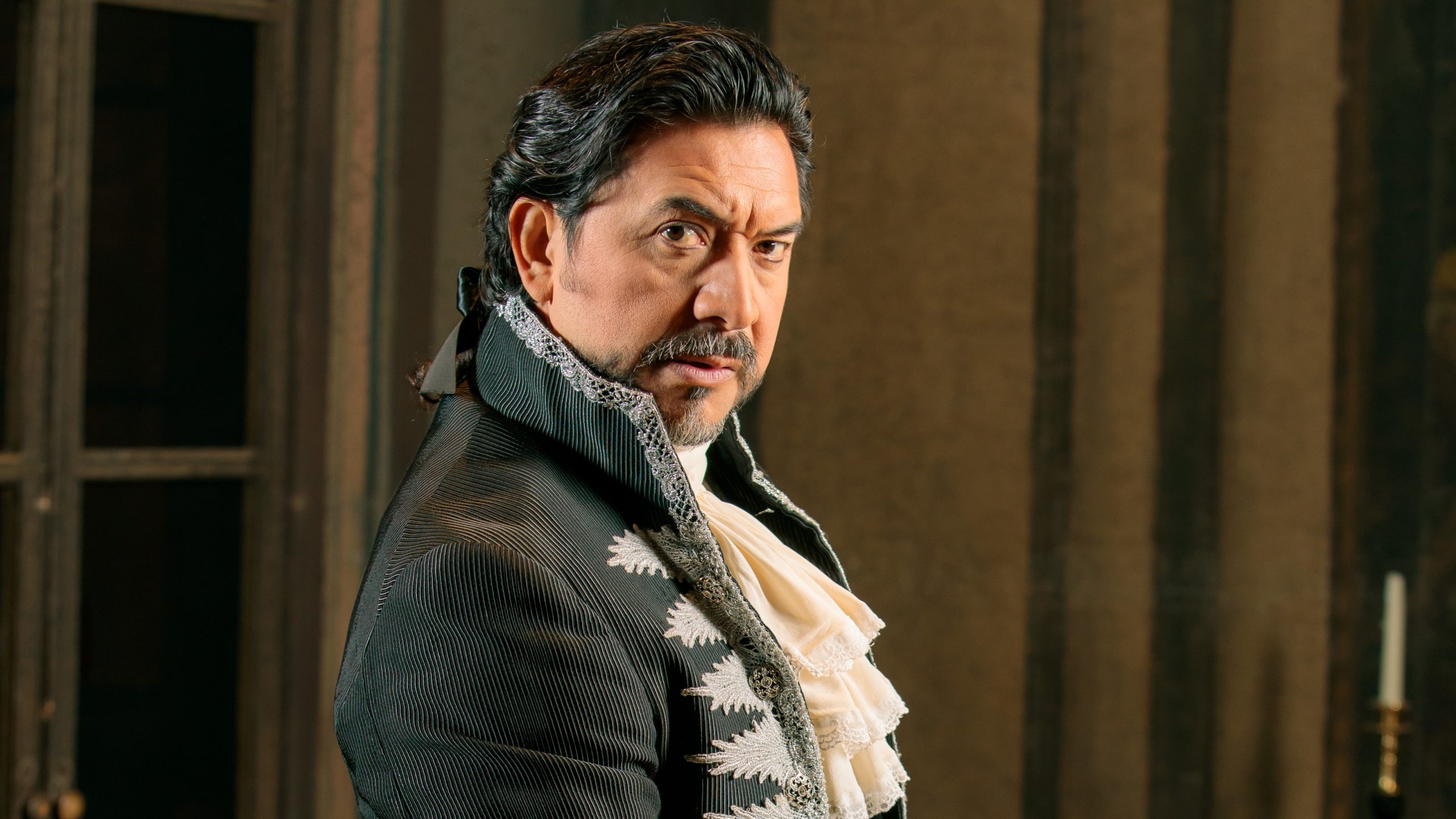 Luis Lesdesma in costume as Baron Scarpia in Tosca smolders at the camera. He is wearing an ornate black top coat and his shiny black hair is pulled back with a black ribbon.