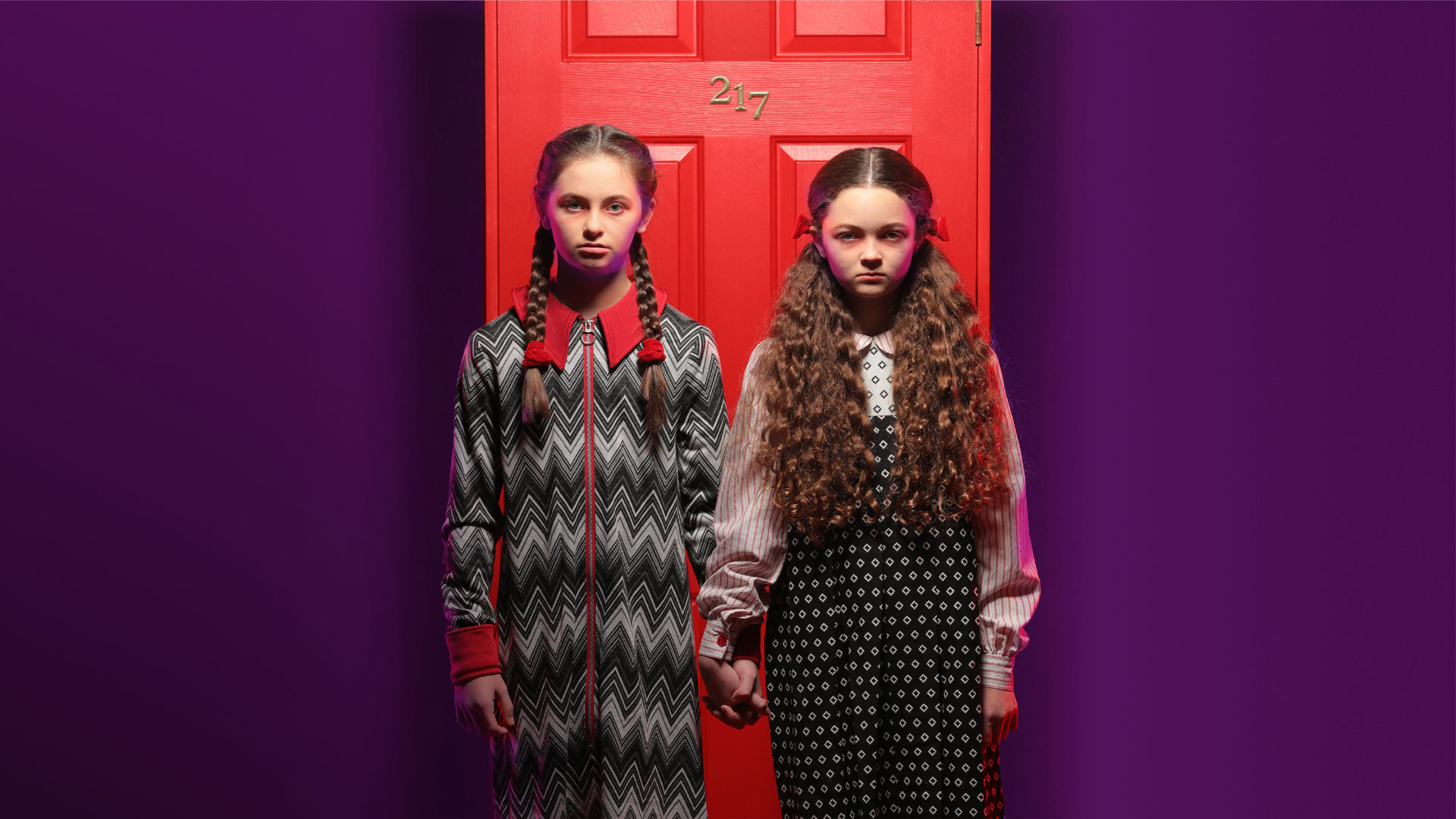 Two grim sisters stand hand in hand in front of a bright red door labeled 217.