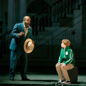 Dick Hallorann wearing a blue suit, sings to Danny who is seated on a suitcase.