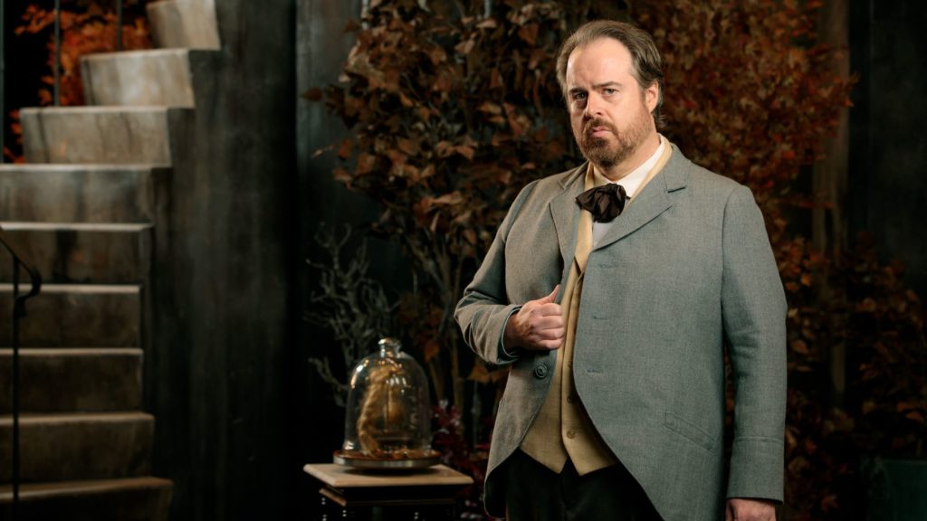 A man stands in front of a bell jar looking solemn