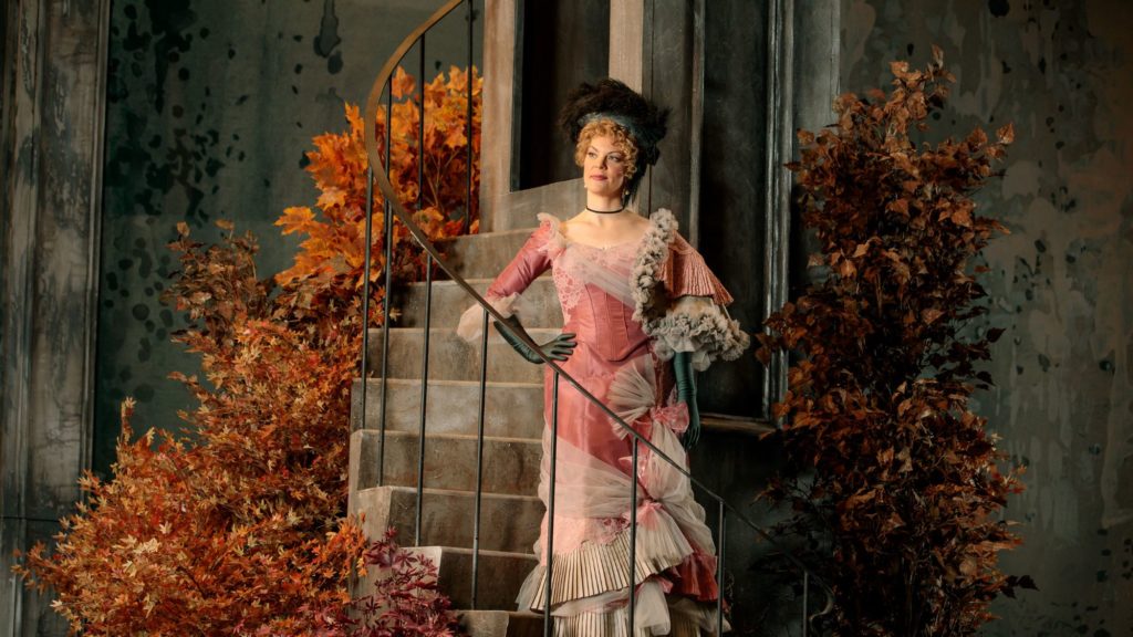 A woman in a pink dress stands on a staircase wuith foliage behind her