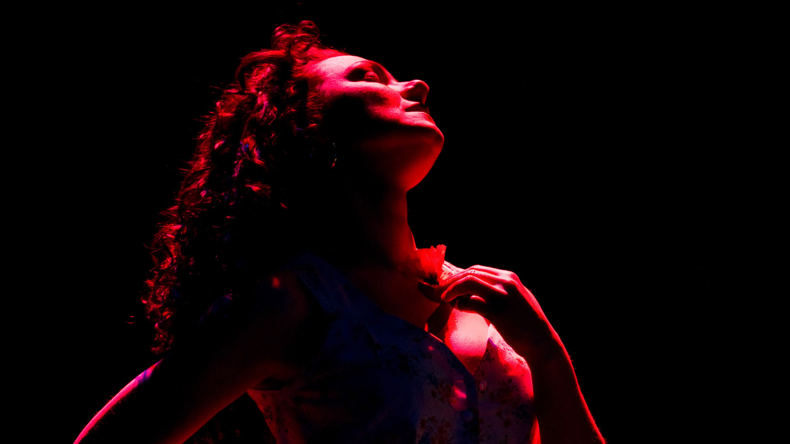 Sandra Eddy as Carmen stands in silhouette holding a red flower with red lighting her from behind. 