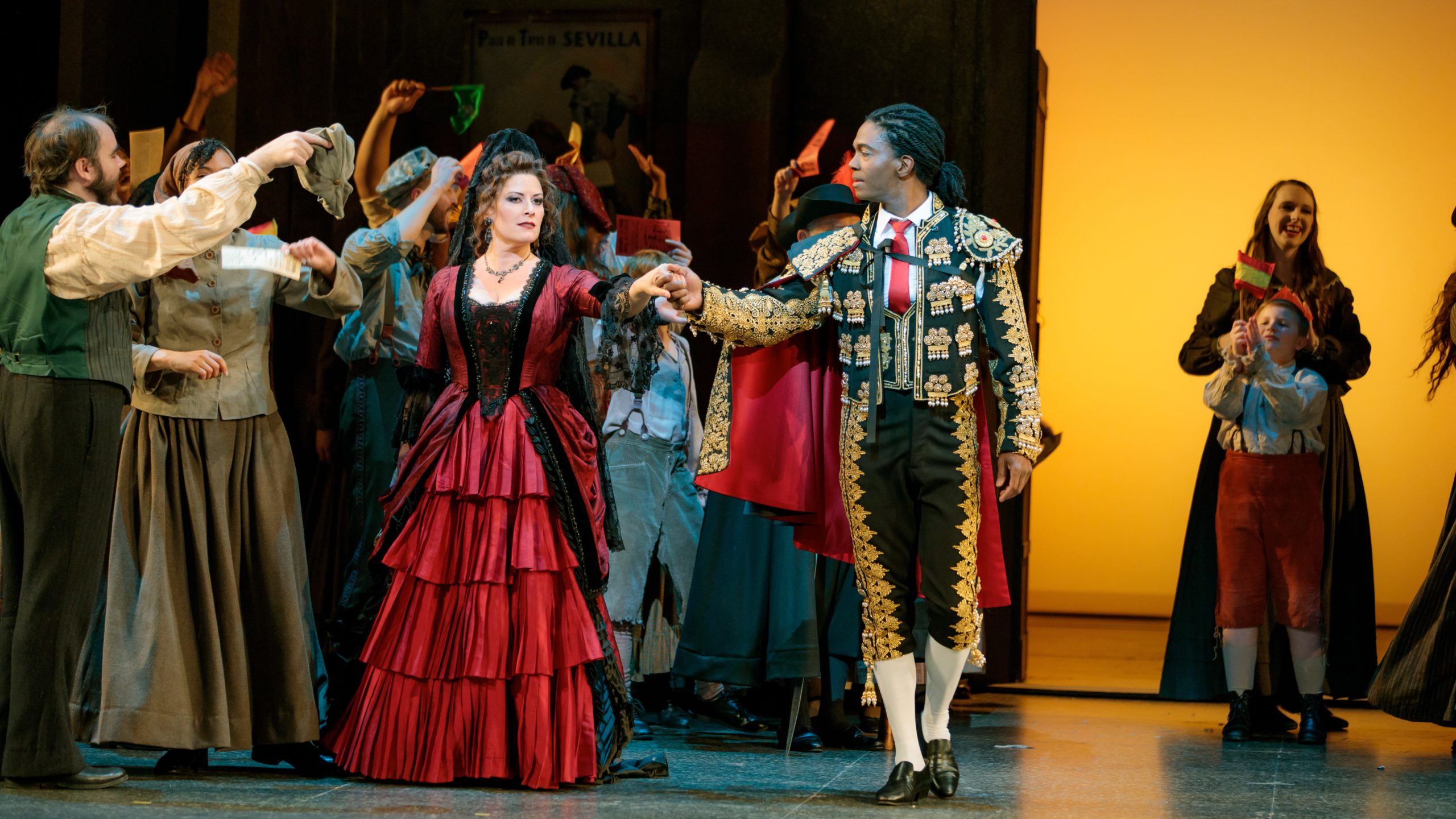 A woman in a long red dress and a man in a black suit with gold decorations and a red cape and tie hold hands as they walk by a cheering crowd. 