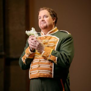 A man with a green and golden military coat gazes lovingly at a white flower he is holding with both hands in front of his body.