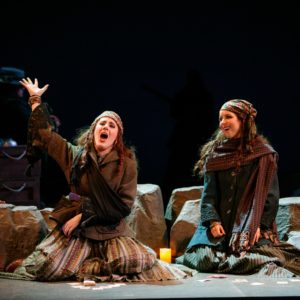 Two women kneel on the group side by side with cards arranged in front of them. The woman on the left has her right arm raised above her head as she sings.