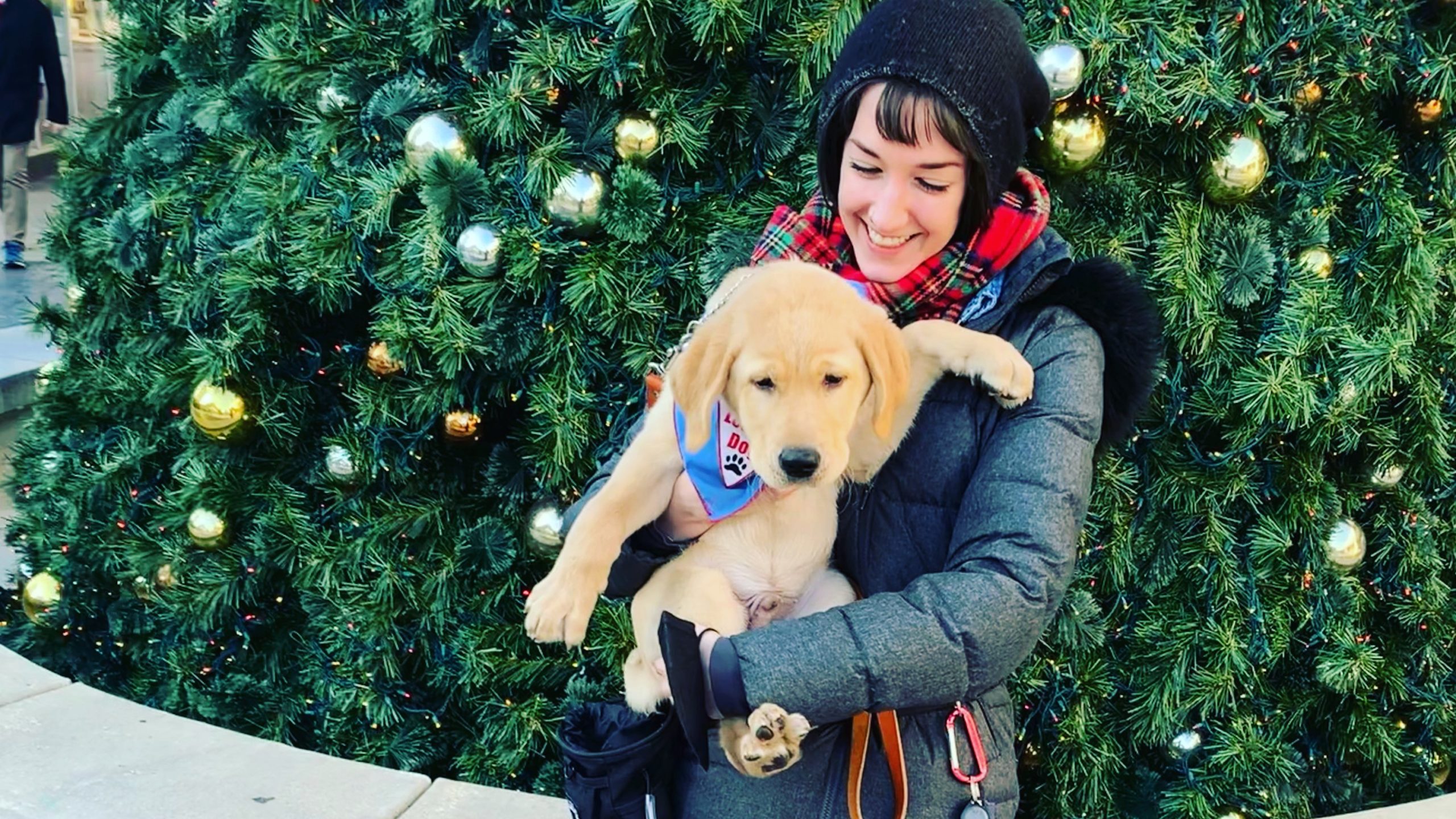 A woman in a black coat and red scarf stands in front of an evergreen tree holding a golden-furred puppy.