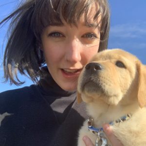 Closeup of a woman with short black hair and bangs holding her golden-furred puppy close to her face.