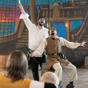 Three performers stand in front of a backdrop with a painted pirate ship in the background. Two have their arms spread wide as they sing, and one stands to the side. The backs of the audiences’ heads are in the bottom left corner. 