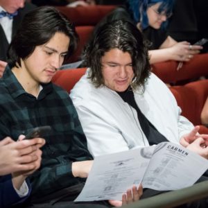 Two students sit in the audience. Two are looking at the performance’s program book.