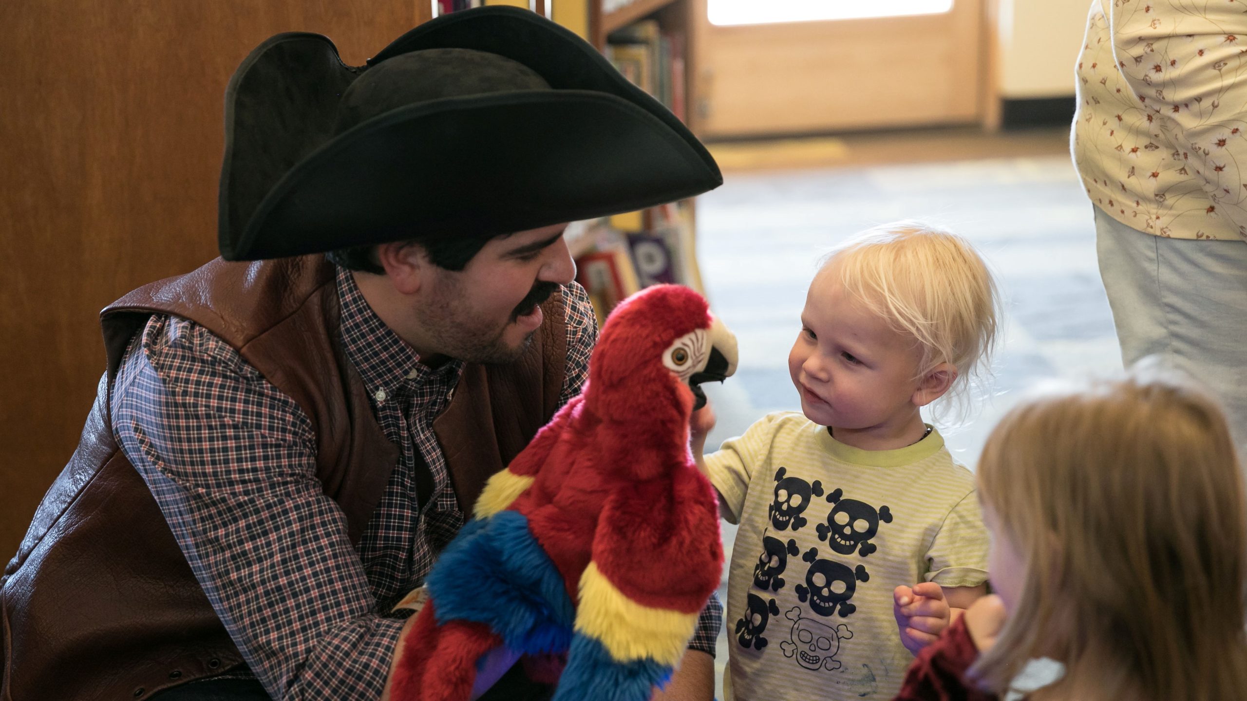 A little boy stands on the right and looks in awe at a red parrot puppet being held by one of our Artists in Residence wearing a three-corner pirate hat. 