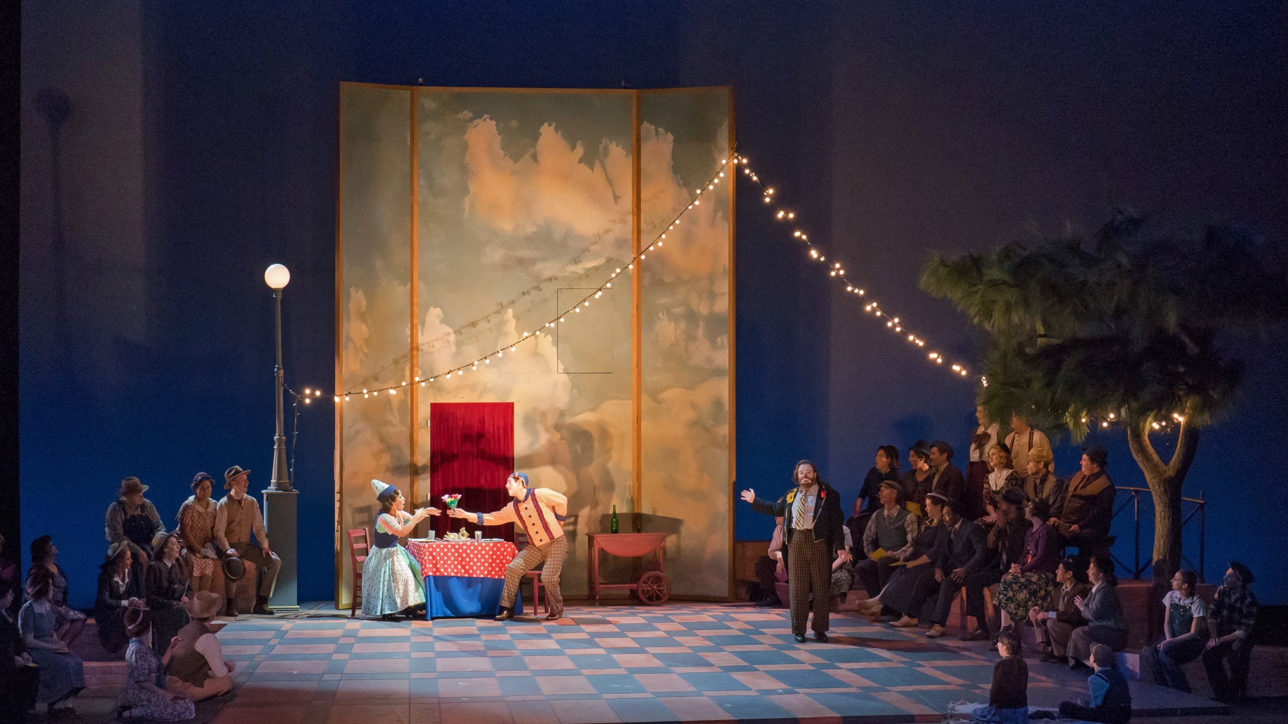 A softly lit stage shows a crowd watching a troupe of actors in clown makeup perform.