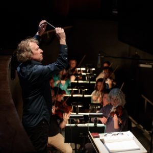 Conductor Ari Pelto stands with his hands raised in an orchestra pit. The violin section is visible behind him.