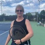 A woman with a black shirt stands on a tennis court holding a racket with her left arm across her body.