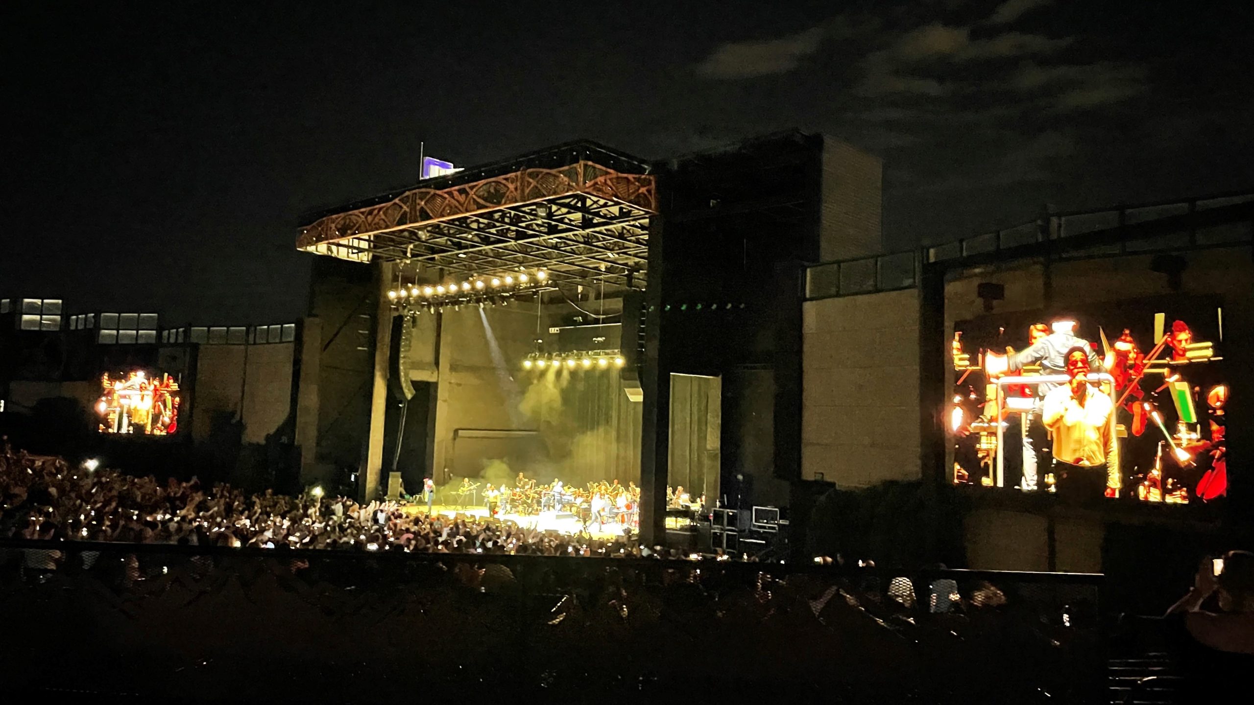 A stage illuminated with yellow light against a night sky in the middle with a large crowd of people in front.