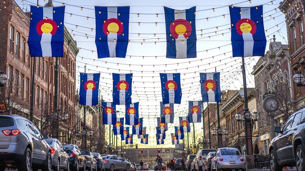 Colorado flags (blue-white-blue stripes with a red “C” with a yellow center) hand vertically above a street with tan buildings on both sides and cars lining the edges.