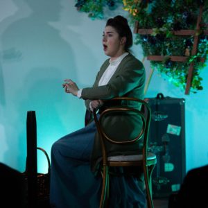 Kira Dills-DeSurra sits facing the right, in costume as Gertrude Stein.