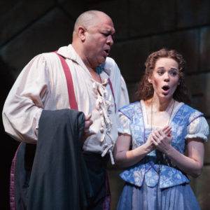 A woman wearing a blue dress holds her hands in front of her body and sings as an older man in a white shirt looks over her. 