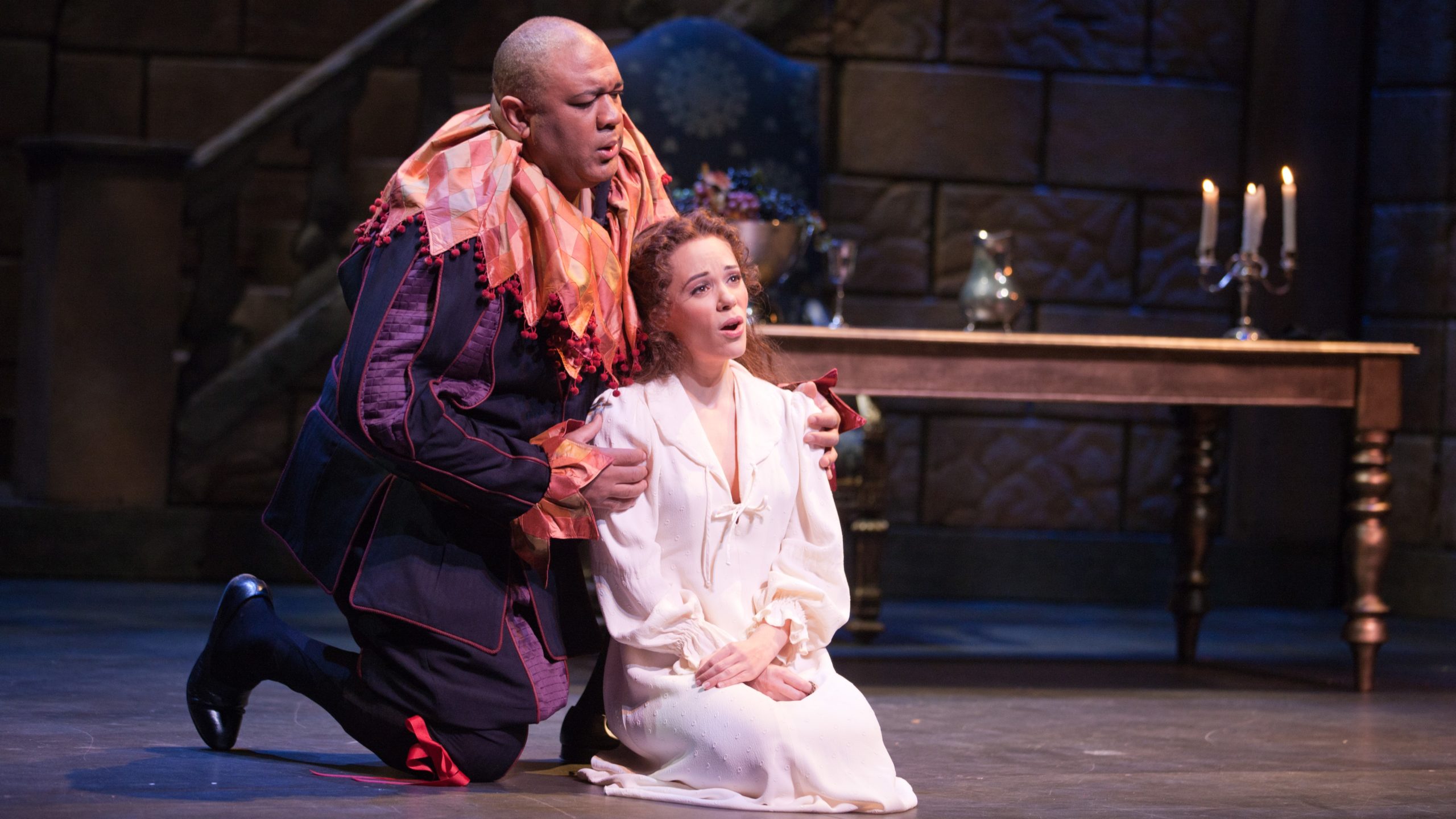 An older man dressed as a jester and his daughter dressed in a white full length shift kneel and embrace each other while singing.