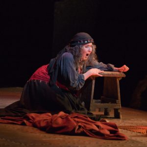 A woman wearing a black head scarf and red cloth around her waist, both embellished with coins, along with a flowing black dress, kneels over a bench in anguish.