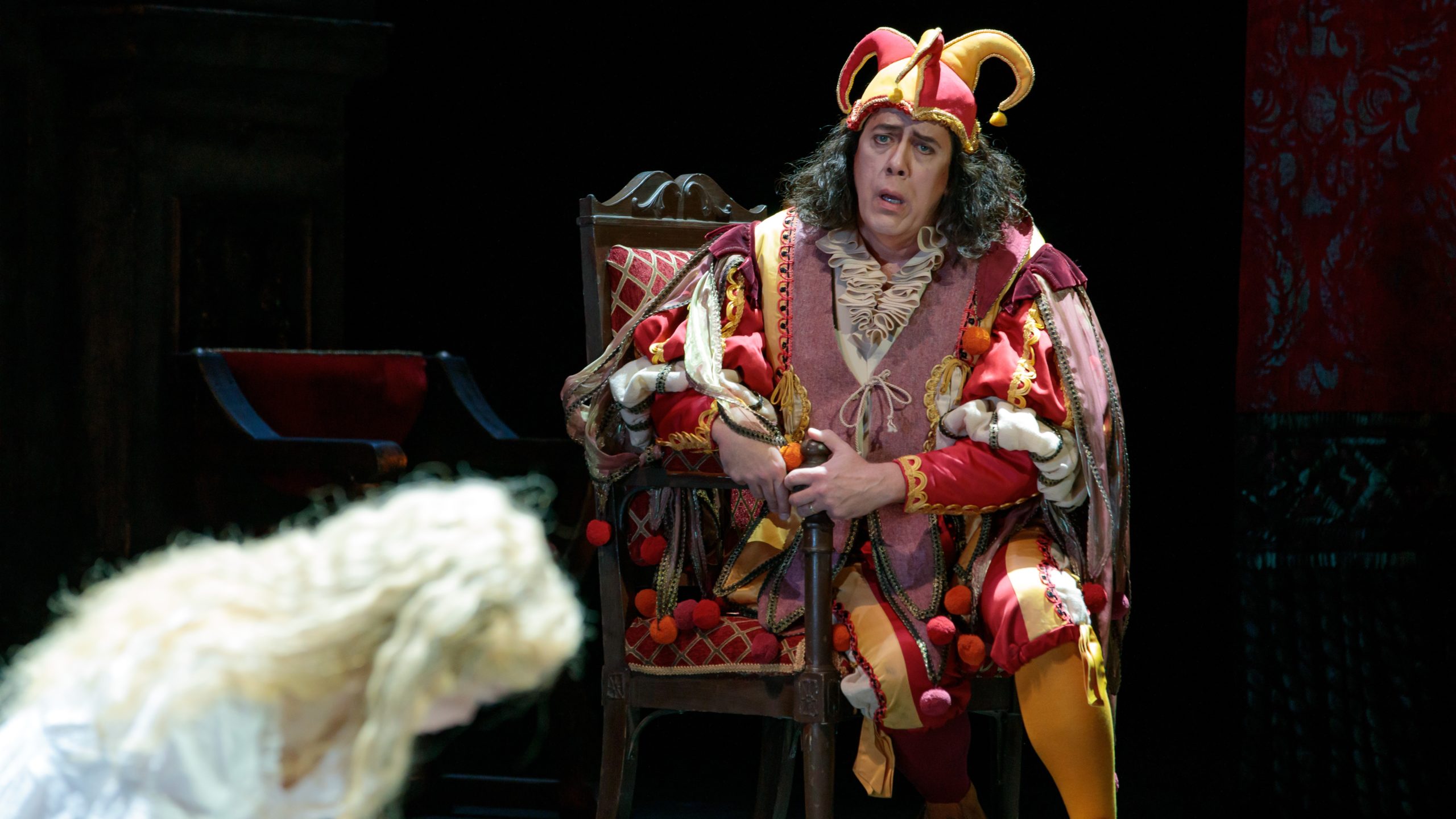 Gilda, a blonde woman is bent over crying in the out-of-focus foreground, while Stephen Powell as Rigoletto in jester's garb sitting looking at his daughter from above.