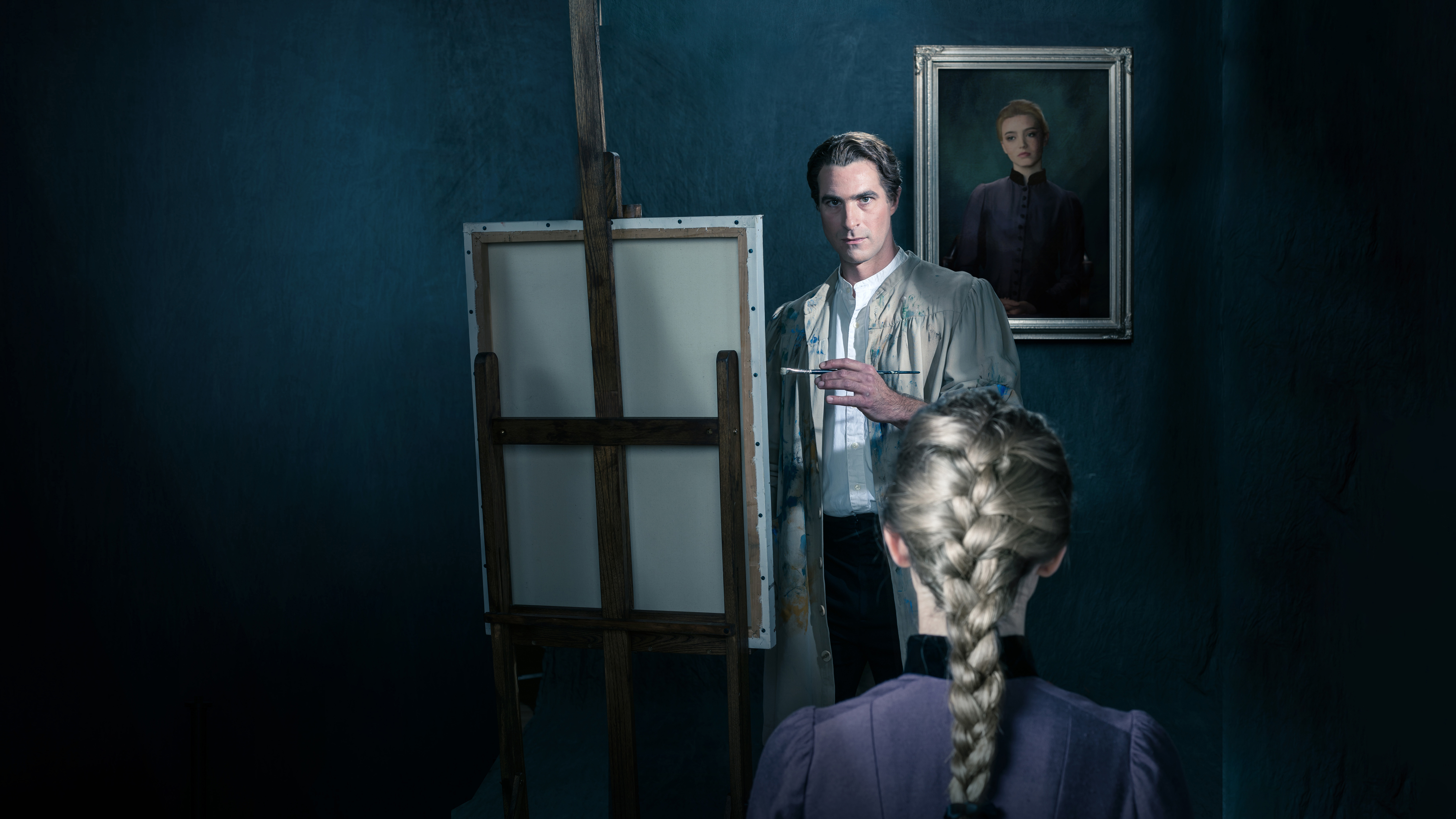 A man stands in front of an easel painting a woman in front of him with a long, blonde braid. A photo of the same woman in a picture frame sits behind the painter.