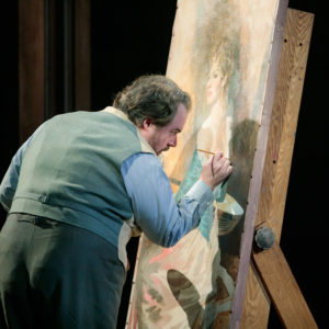 A man holds a paintbrush in front of a painting on canvas.