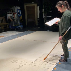 A woman using a long stick with charcoal on the end to sketch a building.