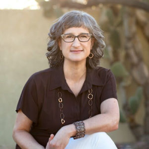 A woman with gray hair and glasses sits and looks at the camera