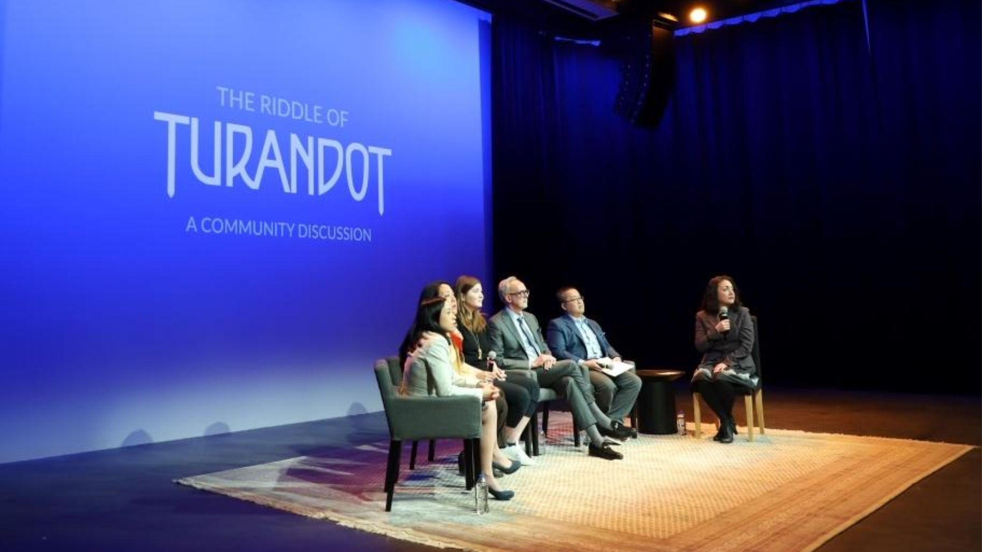 Six people sit in a row on a stage in front of a blue background with text saying "The Riddle of Turandot: A Community Discussion