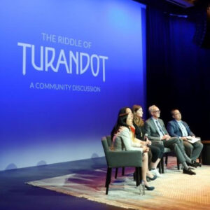 A row of people sit in front of a screen which reads "The Riddle of Turandot: A Community Discussion"