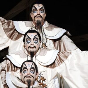 Three men with exaggerated makeup and mustaches stand in a line