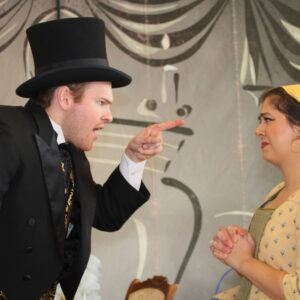 A man in a top hat points his finger harshly at a woman in a yellow dress