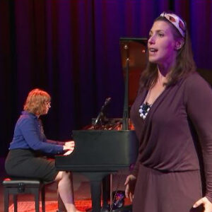 A woman in blue plays the piano while another woman in purple stands in front singing