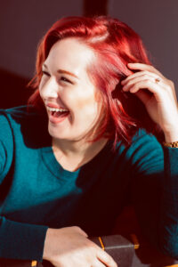 A woman with red hair and a teal shirt smiles away from the camera
