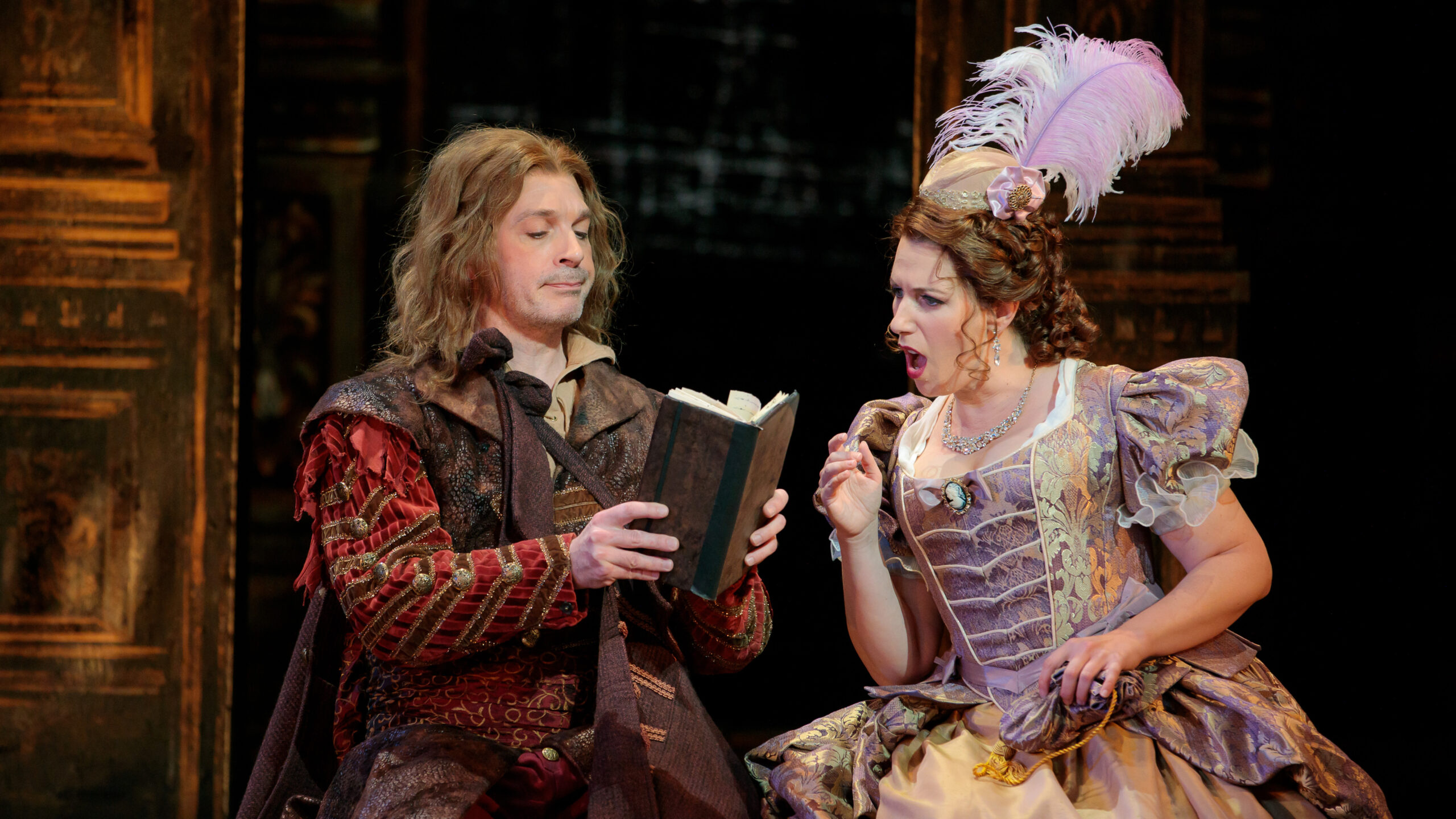 A man holds out a book for a woman to see and she looks on with a shocked face