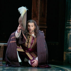 A man in a cloak holds up a hat with a white feather