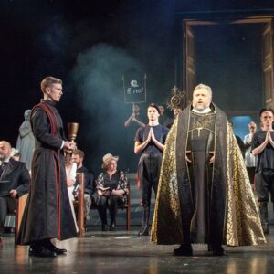 A man in a black and gold cape faces the audience while another man in a black and red cape stands beside him