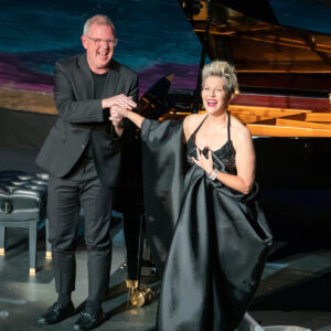A man in a black suit stands in front of a piano and holds hands with a woman in a long black dress