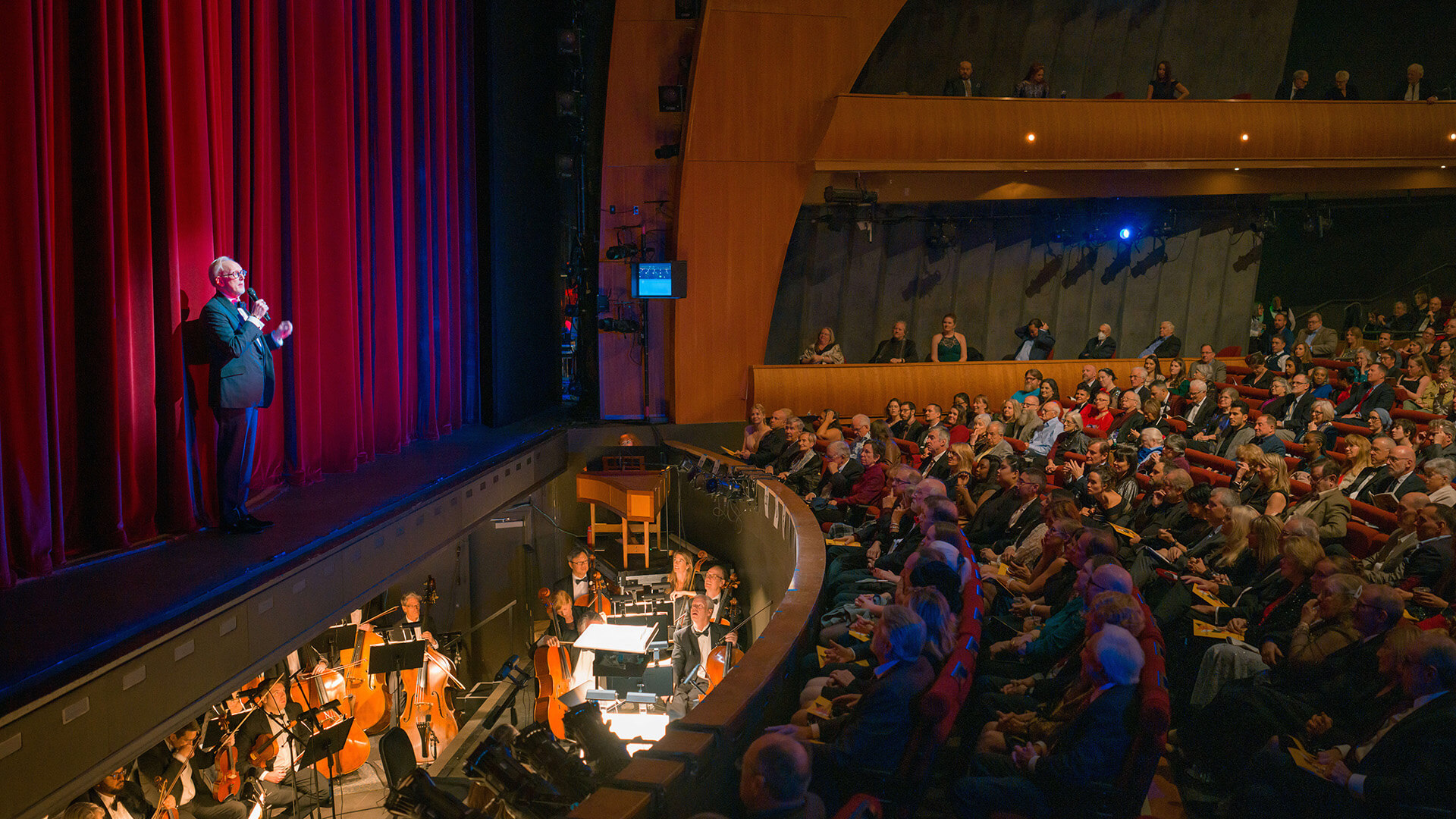 A man in a suit stands on stage in front of a red curtain and looks out to a crowded audience.