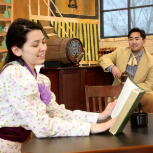 A woman reads a book while a man looks at her longingly