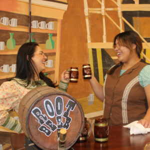 Two women cheers with large cups in front of a barrel that says "Root Beer"