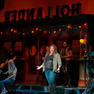 A woman sings in front of a storefront with string lights