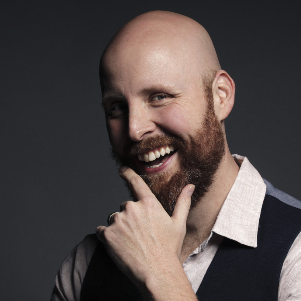 A headshot of a man with a brown beard smiles at the camera