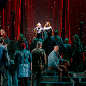 A man and woman stand above a crowd with a red curtain behind them as snow falls