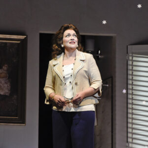 A woman with a brown haired wig and beige blazer jacket stands in front of a window