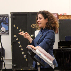 A woman with curly hair points to the crowd while standing in front of a music stand