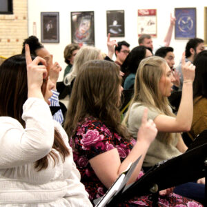 A group of people sit in chairs and sing with their hands pointing upward